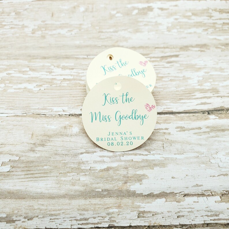May include: A stack of round, cream-colored tags with a hole at the top. Each tag has the text "Kiss the Miss Goodbye" in teal script, with a pink glitter heart. The bottom of the tag reads "Jenna's Bridal Shower 08.02.20".