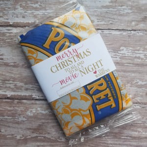 May include: A packaged bag of popcorn with a label that reads "merry CHRISTMAS TO ALL AND TO ALL A movie NIGHT". The popcorn bag has blue and yellow branding. The package is sealed in clear plastic.
