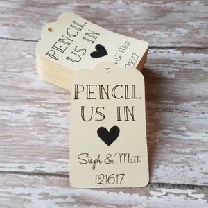 Op de afbeelding: Een stapel cr&egrave;mekleurige labels met een geschulpte bovenrand. Elk label heeft de zwarte tekst "PENCIL US IN", een zwart hart en de namen "Steph & Matt" met de datum "12.16.17".
