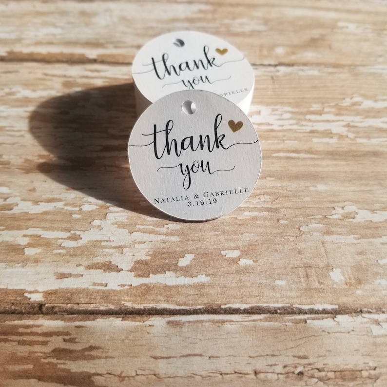 May include: White, round tags with the words "thank you" in black script and a gold heart. The tags also include the names "Natalia & Gabrielle" and the date "3.16.19".