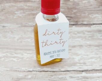 Könnte beinhalten: Kleine Glasflasche mit rotem Verschluss, gefüllt mit einer goldenen Flüssigkeit. Ein weißes Etikett ist angebracht, mit den Worten "dirty thirty" in kupferfarbener Schrift, zusammen mit "Ashley's 30th Birthday May 1, 2022".