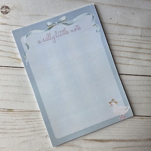 May include: A light blue notepad with a decorative border and the text "a silly little note" in cursive. A cartoon duck wearing a hat is in the lower right corner. The notepad has a light blue ribbon design at the top.