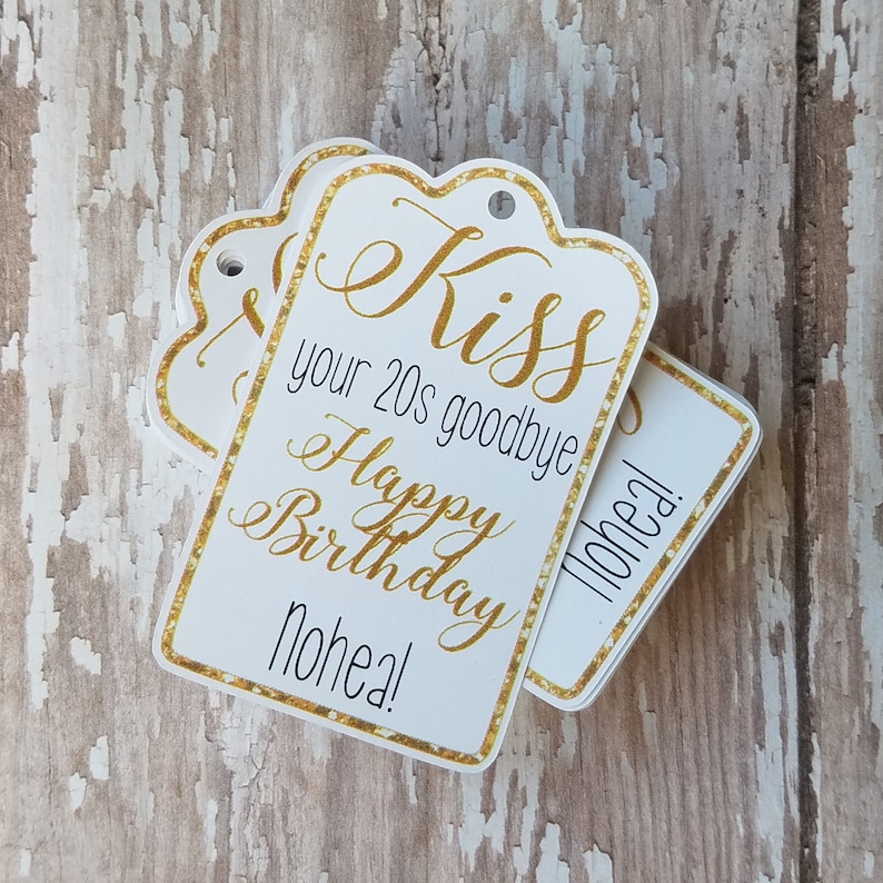 May include: White gift tags with gold trim and text that reads "Kiss your 20s goodbye Happy Birthday Nohea!" in gold script. The tags are stacked on a weathered wood surface.