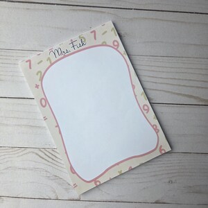 May include: A rectangular notepad with a white interior and a pink border. The border is decorated with numbers and symbols. The name "Mrs. Fish" is written in cursive at the top.