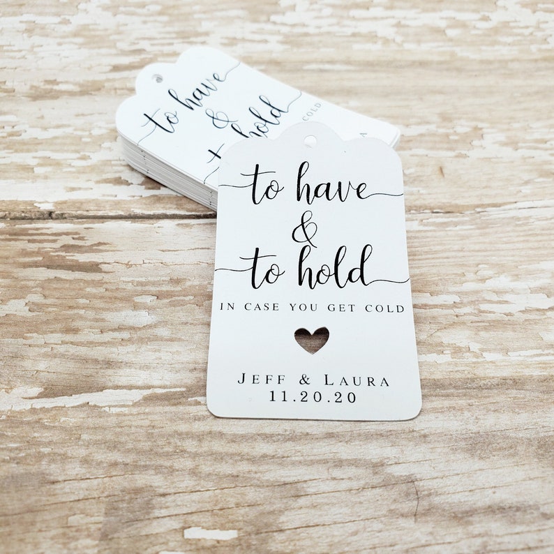 May include: White gift tags with a scalloped top edge, stacked on a wooden surface. The tags read "to have & to hold" with a heart cutout and the text "IN CASE YOU GET COLD". Names and a date are printed at the bottom.