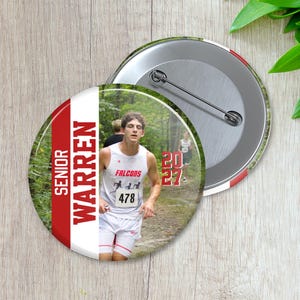 Op de afbeelding: Een ronde pinback-button met een foto van een hardloper in een wit hemd met de tekst FALCONS en 478. Het ontwerp bevat een rode verticale streep met de tekst SENIOR WARREN en het jaartal 2027 in het rood aan de rechterkant.