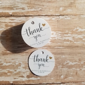 May include: Two white, round gift tags with the words "thank you" in elegant black script. Each tag has a small gold heart and the names "Natalia & Gabrielle" with the date 3.16.19.