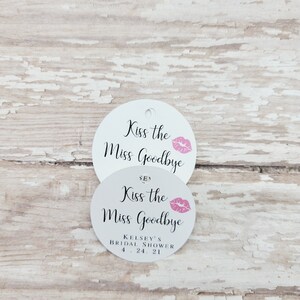 May include: Two white, round tags with the text "Kiss the Miss Goodbye" in black script and a pink lip graphic. The bottom tag includes "Kelsey's Bridal Shower 4.24.21". One tag has a hole for hanging.
