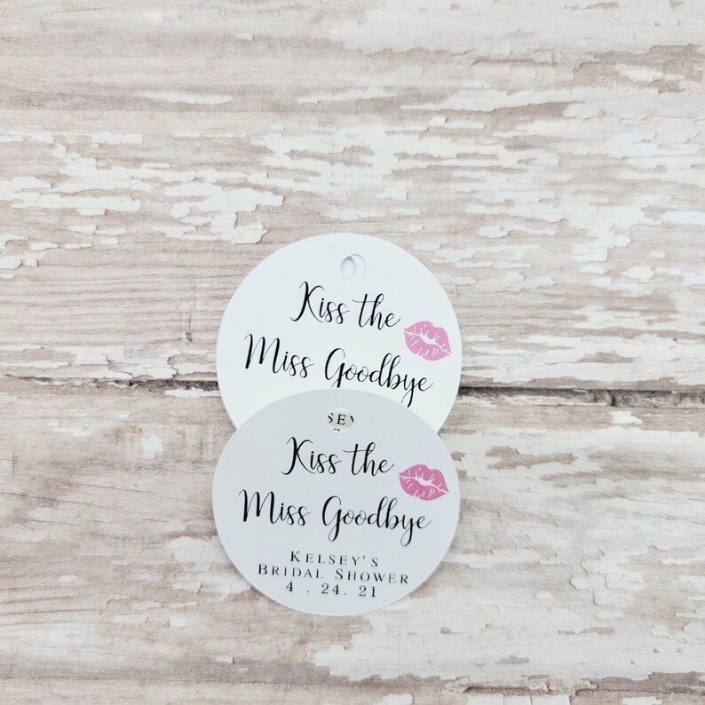 May include: Two white, round tags with the text "Kiss the Miss Goodbye" in black script and a pink lip graphic. The bottom tag includes "Kelsey's Bridal Shower 4.24.21". One tag has a hole for hanging.