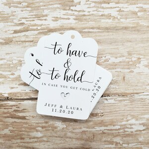 May include: White wedding favor tags with a scalloped top edge. The tags have the text "to have & to hold" in elegant script, along with "IN CASE YOU GET COLD" and the names "JEFF & LAURA" and the date "11.20.20". A small heart cutout is visible.
