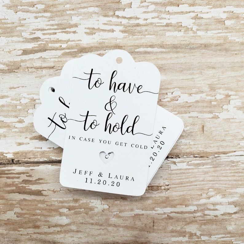 May include: White wedding favor tags with a scalloped top edge. The tags have the text "to have & to hold" in elegant script, along with "IN CASE YOU GET COLD" and the names "JEFF & LAURA" and the date "11.20.20". A small heart cutout is visible.