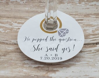 May include: A white, round wine glass charm with the text "He popped the question... She said yes!" and the date 7.20.2019. The charm features a gold ring design and a gray diamond illustration.