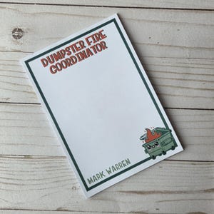 Könnte beinhalten: Ein weißer Notizblock mit grünem Rand und dem Text "DUMPSTER FIRE COORDINATOR" in Rot. In der Ecke befindet sich ein Cartoon-Müllcontainer mit Flammen. Der Name "MARK WARREN" ist unten aufgedruckt.