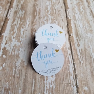 May include: A stack of white, round gift tags with a light blue "thank you" script and a gold heart. The tags also include the text "CHRIS AND GLORIA'S BABY SHOWER 5.11.19". Each tag has a small hole at the top.
