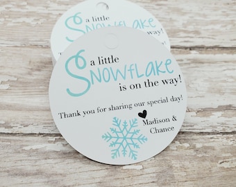 以下が含まれることがあります： 水色の雪の結晶のデザインと「a little SNOWFLAKE is on the way!」の文字が入った、白い丸いタグが2つ。タグには「Thank you for sharing our special day!」の文字と「Madison & Chance」の名前も含まれています。