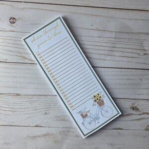May include: A rectangular notepad with a green border and the text "shine through your to-dos" in gold script. The notepad features a bicycle illustration with sunflowers and a basket. The notepad has lined spaces for a to-do list.
