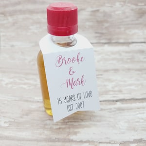 Könnte beinhalten: Kleine Glasflasche mit rotem Verschluss, gefüllt mit einer goldenen Flüssigkeit. Ein weißes Etikett ist angebracht, mit den Namen "Brooke & Mark" in rosa Schrift und "15 YEARS OF LOVE EST. 2007" in schwarzem Text.