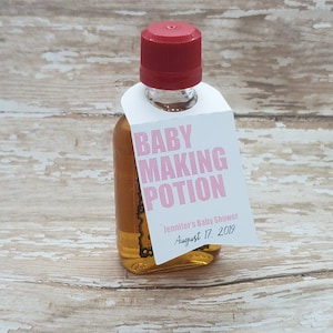 Może przedstawiać: Mała szklana butelka z czerwoną zakrętką, wypełniona złotym płynem. Dołączona biała etykieta z różowym napisem "BABY MAKING POTION", a także "Jennifer's Baby Shower" i data 17 sierpnia 2019.