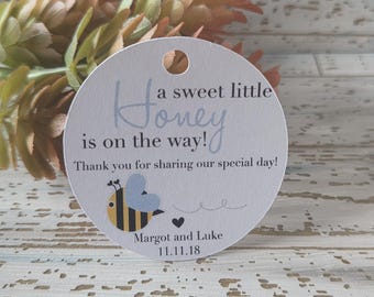 Op de afbeelding: Een ronde, lichtgrijze tag met een gat aan de bovenkant. De tag bevat de tekst "A sweet little Honey is on the way!" in lichtblauwe letters. Een cartoonbij en de namen "Margot en Luke" staan ook op de tag.