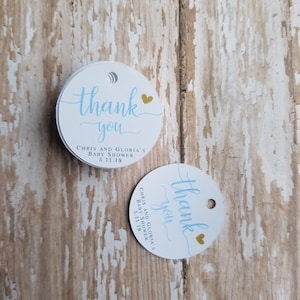 May include: Two white, round thank you tags with a light blue script font. The text "thank you" is above the text "CHRIS AND GLORIA'S BABY SHOWER 5.11.19". A small gold heart is to the right of the words "thank you".