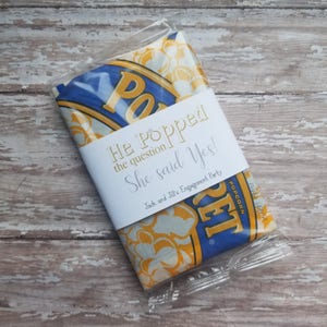 May include: A clear cellophane-wrapped bag of popcorn with a white label. The label reads "He Popped the question She said Yes!" in gold and silver lettering. The popcorn bag has a blue and yellow design with the word "POPCORN" visible.