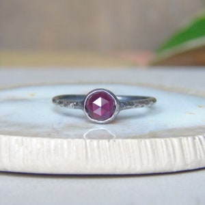 Könnte beinhalten: Ein silberner Ring mit einem facettierten, tiefroten Edelstein. Der Ring hat eine gehämmerten Textur am Band und eine Zargenfassung für den Stein. Der Edelstein ist rund und fängt das Licht ein, wodurch seine vielen Facetten zur Geltung kommen.