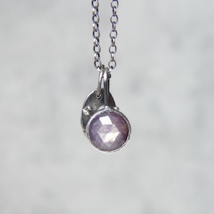 Può includere: Una collana d'argento con un piccolo pendente di pietra preziosa sfaccettata viola chiaro. Il pendente è rotondo e incastonato in una lunetta d'argento, con un piccolo ciondolo a forma di foglia. La catena è delicata e argentata.