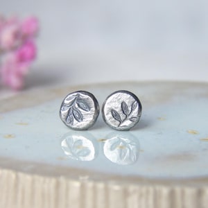 Puede incluir: Un par de pendientes de botón plateados. Cada pendiente presenta un diseño circular con una superficie texturizada y un patrón de hojas detallado. Los pendientes se muestran sobre una superficie clara, reflejando la luz.
