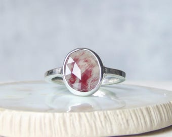 以下が含まれることがあります： ファセットカットが施された楕円形の石をあしらったシルバーリング。石は半透明の白をベースに、深みのある赤色の繊維状または筋状のインクルージョンが全体に広がっており、滑らかな金属製のバンドにセットされています。