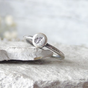 Puede incluir: Un anillo de plata con un pequeño diseño de hoja en la parte superior. El anillo es simple y delicado.