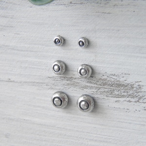 Könnte beinhalten: Sechs silberne Ohrstecker in drei verschiedenen Größen. Die Ohrringe haben ein strukturiertes, gehämmertes Aussehen mit einem kleinen, dunklen Kreis in der Mitte. Die Ohrringe sind in zwei Reihen zu je drei Stück auf einer weißen, verwitterten Holzoberfläche angeordnet.