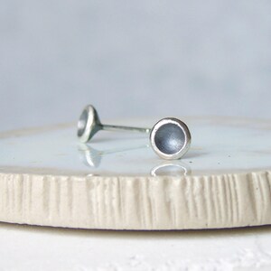 May include: A pair of silver stud earrings. Each earring has a small, circular, concave design on a thin post. The earrings are presented on a light-coloured, textured surface, with a soft, blurred background.