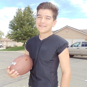 May include: A person wearing a black V-neck shirt and holding a brown football. The person is wearing a yellow and white flag football belt. The background includes a tree, houses, and a blue sky.