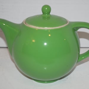 May include: A bright green ceramic teapot with a rounded body, a curved spout, and a loop handle. The lid has a small knob on top and a cream-colored rim. The teapot is designed for brewing tea.