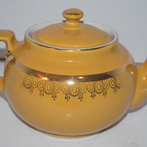 May include: Mustard yellow ceramic teapot with a lid and spout. The teapot has a gold band with a black decorative pattern around the middle. The handle and lid finial are also mustard yellow. A classic tea accessory.