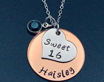 May include: A silver necklace with a copper pendant that says "Sweet 16 Haisley". The pendant has a silver heart charm with the words "Sweet 16" inscribed on it. A dark green gemstone dangles from the chain.