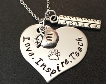 Op de afbeelding: Zilveren hartvormige ketting met de woorden "Love. Inspire. Teach." erop gegraveerd. De ketting heeft een klein appelbedeltje, een pootbedeltje en een liniaalbedeltje.