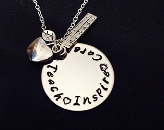 May include: Silver necklace with a round pendant inscribed with "Teach, Care, Inspire." The necklace has a heart charm, a leaf charm, and a small ruler charm. The chain is fine and the background is black.