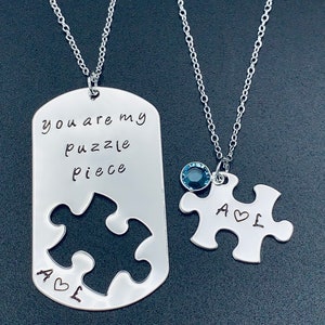 May include: Two silver necklaces with puzzle piece pendants. One pendant has the words "you are my puzzle piece" engraved on it and the other has the initials "A & L" with a heart symbol. The necklace with the initials has a small blue gemstone.