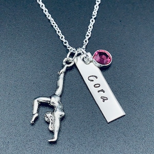 May include: Silver necklace with a gymnast charm, a pink crystal, and a silver tag engraved with the name "Cora".