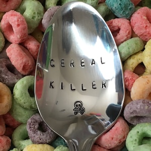 May include: A silver spoon with the words "Cereal Killer" engraved on it, a skull and crossbones symbol is below the text. The spoon is surrounded by colorful cereal rings.