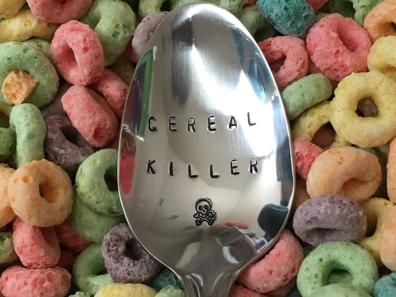 May include: A silver spoon with the words "Cereal Killer" engraved on it, a skull and crossbones symbol is below the text. The spoon is surrounded by colorful cereal rings.