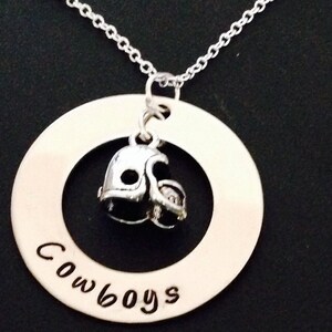 Pode incluir: Colar com pingente prateado com um capacete de futebol dentro de uma moldura circular. A palavra "Cowboys" está inscrita na moldura. O colar pende de uma corrente delicada, perfeito para os fãs de esportes.