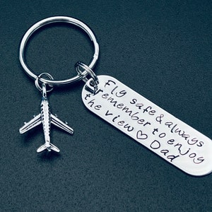 以下が含まれることがあります： 銀色の飛行機のキーホルダー。銀色のタグには「Fly safe & always remember to enjoy the view ♥ Dad」と書いてあります。