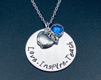 Op de afbeelding: Zilveren ketting met een ronde hanger met de tekst "Love. Inspire. Teach." en een klein zilveren appelbedeltje met een blauwe strasssteen.