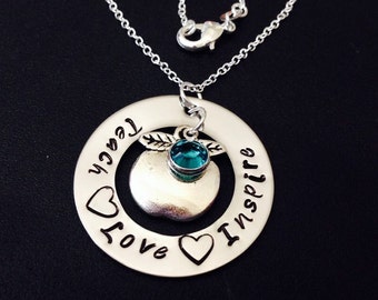 Op de afbeelding: Zilveren ketting met een ronde hanger waarop "Love Teach Inspire" staat met een hart en een appelbedel met een turquoise edelsteen.