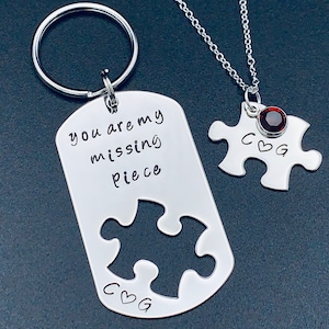 May include: A silver keychain with a dog tag charm that says "you are my missing piece" with a cut-out puzzle piece and the initials "CBG". A silver necklace with a puzzle piece charm with a red gemstone and the initials "CBG".
