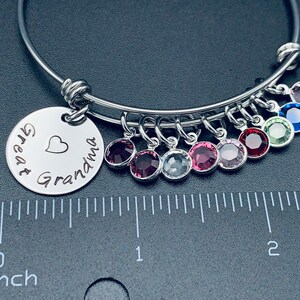 May include: A silver bangle bracelet with a charm that reads "Great Grandma" with a heart. The bracelet has multiple charms with colourful crystals.