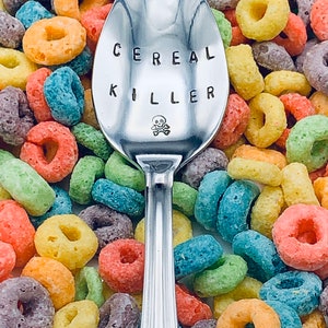 May include: A silver spoon with the words "Cereal Killer" engraved on it, surrounded by colorful cereal rings.