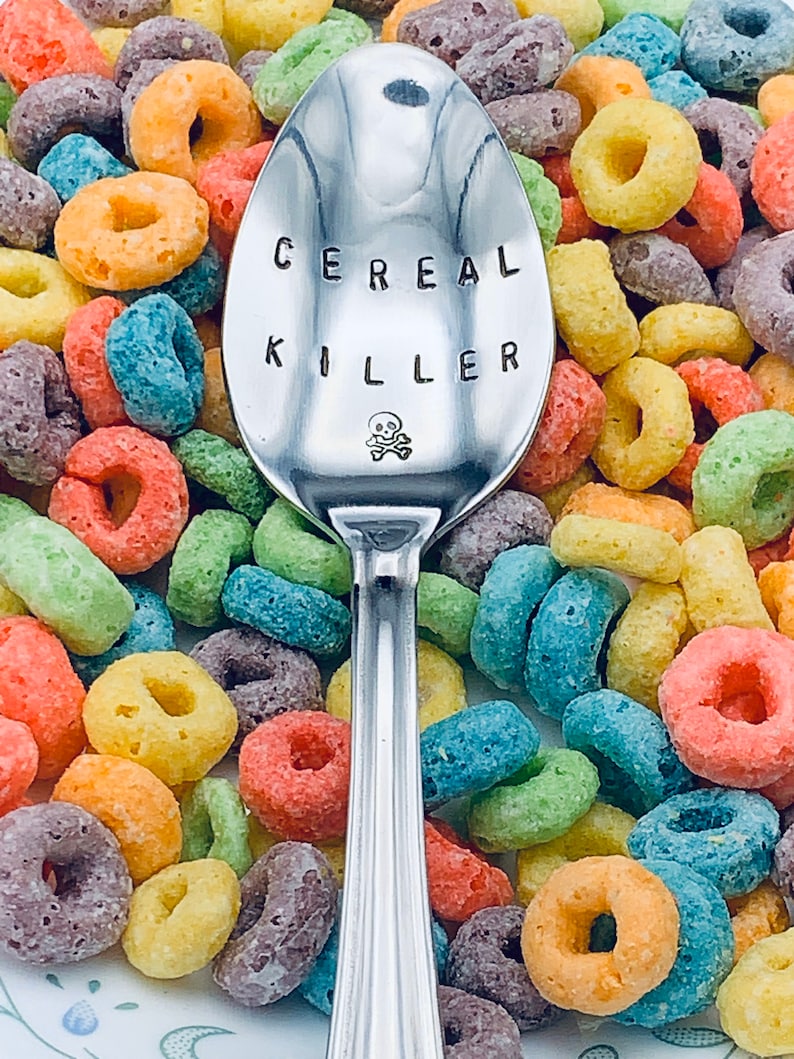 May include: A silver spoon with the words "Cereal Killer" engraved on it, surrounded by colorful cereal rings.